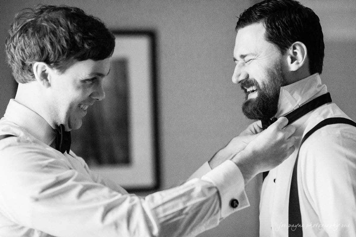 st. mary's chapel raleigh wedding photography - jackie & rob Two men dressed in formal attire sharing a happy moment. One is adjusting the others bow tie with smiles on their faces. The setting appears to be indoor and the image is in black and white.