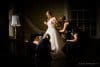 A bride in a white gown stands as four bridesmaids help adjust her dress. The room is dimly lit, with a lamp on the left. The atmosphere is elegant and focused.