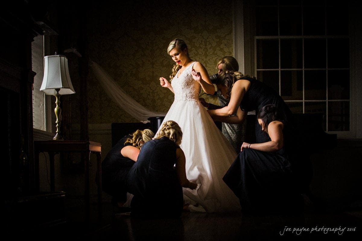 st. mary's chapel raleigh wedding photography - jackie & rob A bride in a white gown stands as four bridesmaids help adjust her dress. The room is dimly lit, with a lamp on the left. The atmosphere is elegant and focused.