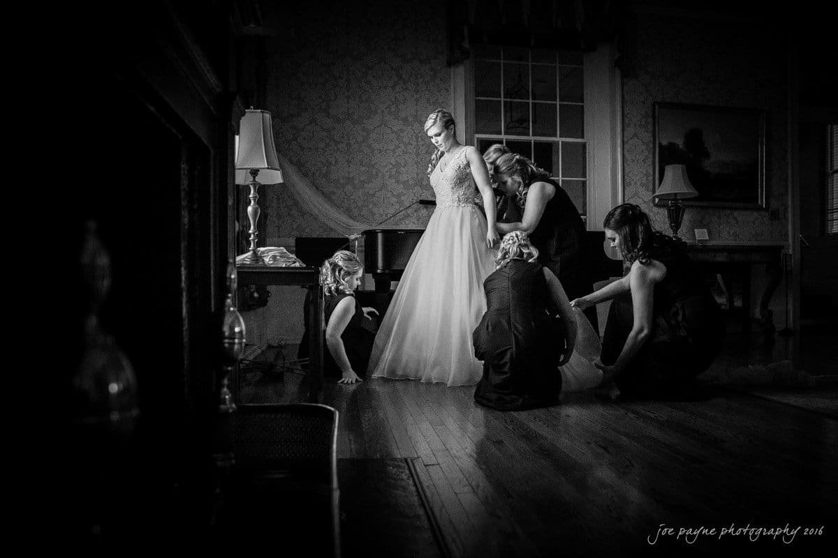 st. mary's chapel raleigh wedding photography - jackie & rob A bride in a long gown stands in a dimly lit room, surrounded by four women helping with her dress. The scene conveys an intimate, pre-wedding moment with a focus on the bride in her detailed attire. Black and white photography enhances the mood.