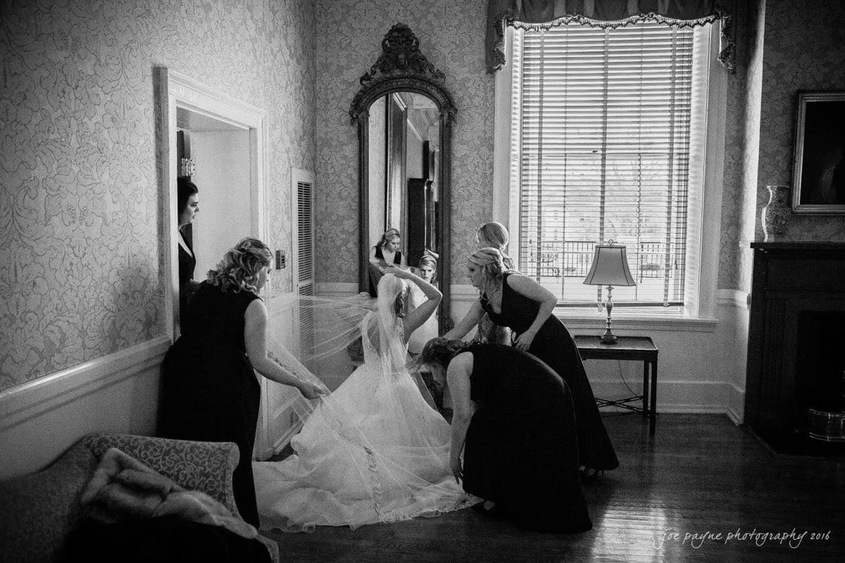st. mary's chapel raleigh wedding photography - jackie & rob A bride in a wedding gown adjusts her veil with the help of her bridesmaids in a vintage-style room. A large mirror and window with curtains adorn the walls. The atmosphere is intimate and elegant.