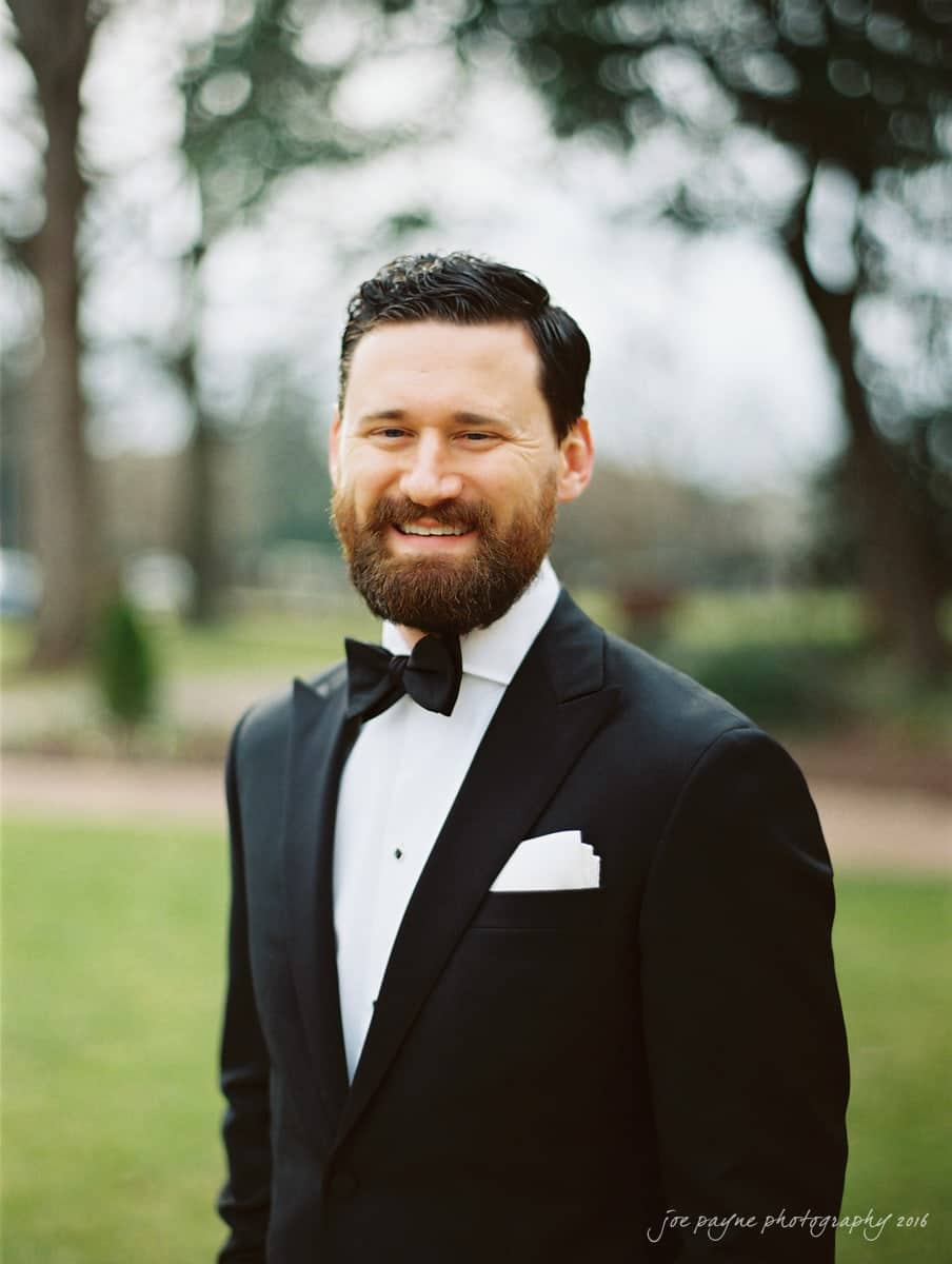 st. mary's chapel raleigh wedding photography - jackie & rob A man with a beard and mustache is wearing a black tuxedo with a black bow tie and a white pocket square. He stands outdoors on a grassy area with blurred trees in the background, smiling at the camera.