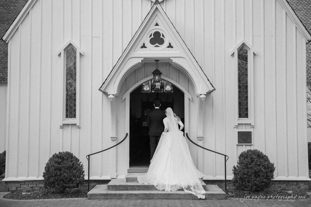 st. mary's chapel raleigh wedding photography - jackie & rob A bride in a flowing white gown and veil walks arm in arm with a groom towards the entrance of a small, white chapel. The architecture features pointed arches and decorative elements. Two small shrubs frame the doorway.