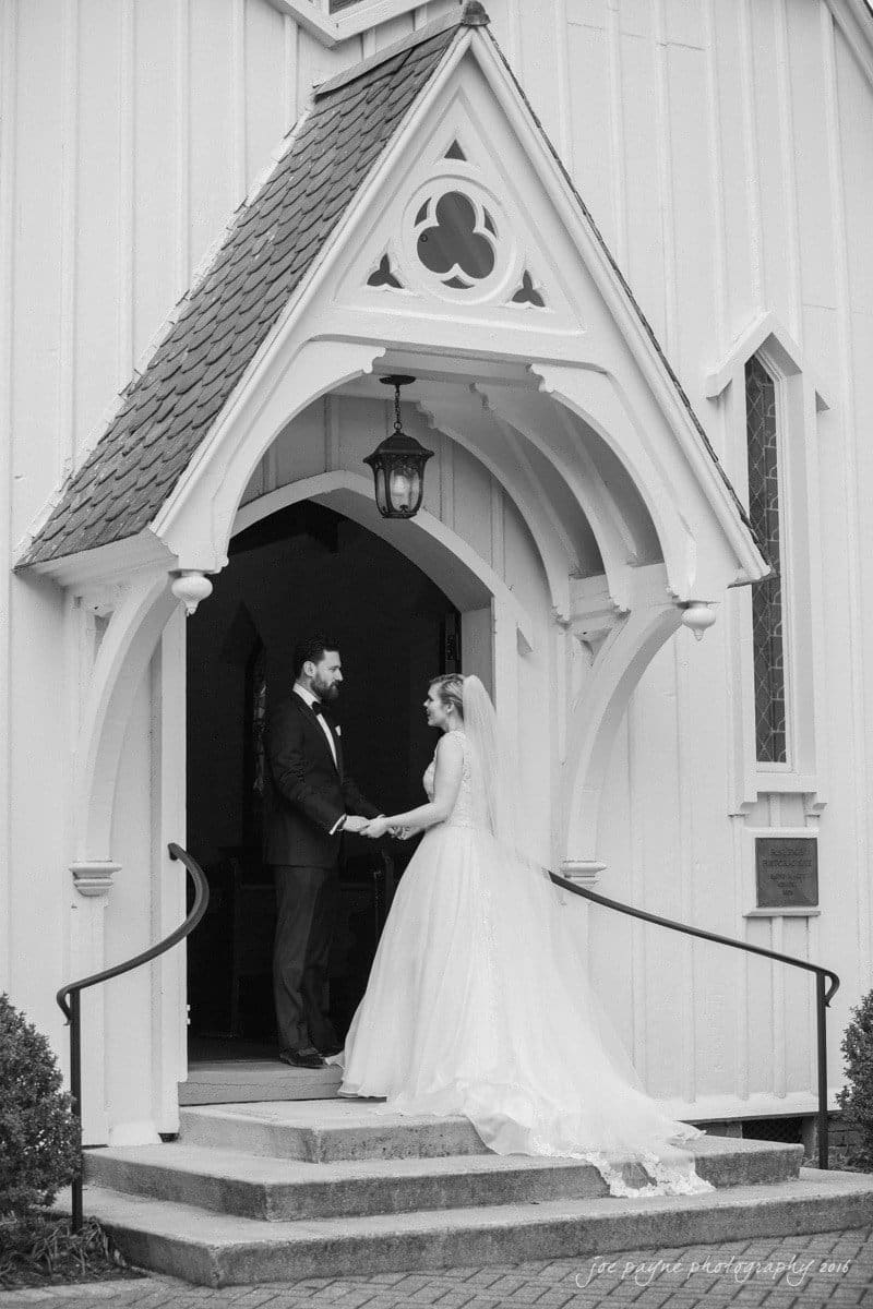 st. mary's chapel raleigh wedding photography - jackie & rob A bride and groom stand holding hands at the entrance of a small, ornate chapel. The bride wears a long, flowing gown and veil, and the groom is dressed in a formal suit. The scene is captured in black and white.
