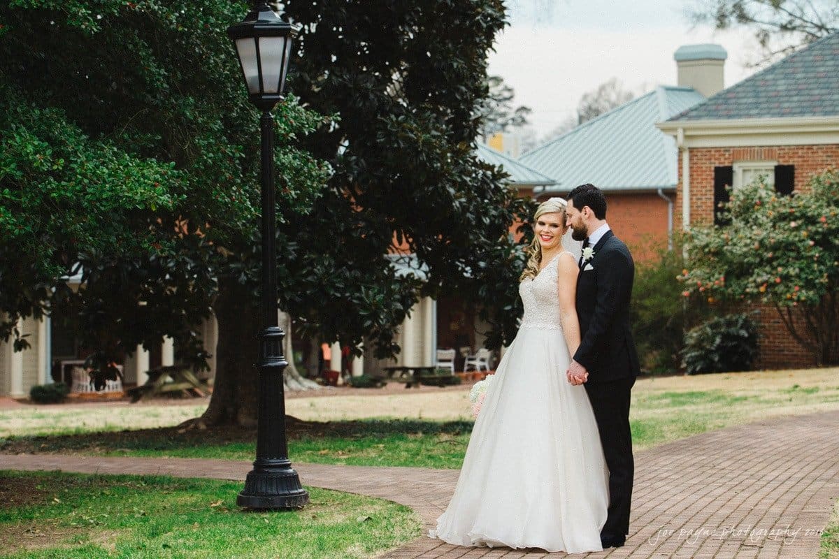 st. mary's chapel raleigh wedding photography - jackie & rob A bride and groom stand on a brick path holding hands, with a historic-style building and a large tree in the background. The bride is in a white gown and the groom is in a black suit. A lamppost is visible beside them.