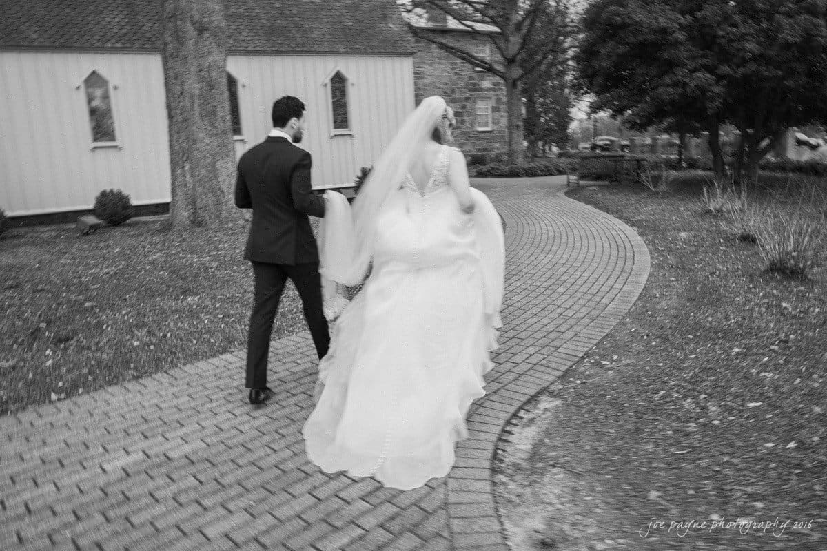st. mary's chapel raleigh wedding photography - jackie & rob A black and white photo of a bride and groom walking away on a brick pathway. The bride is wearing a long gown and veil, and the groom is in a dark suit. They are outside near a chapel or small church. Trees and buildings are in the background.