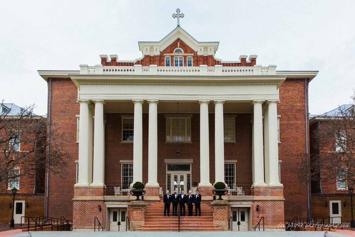 st. mary's chapel raleigh wedding photography - jackie & rob A stately brick building with a grand entrance featuring eight tall white columns. Six people are posed on the steps in front, dressed in formal attire. The architecture includes a prominent cross on the roof.