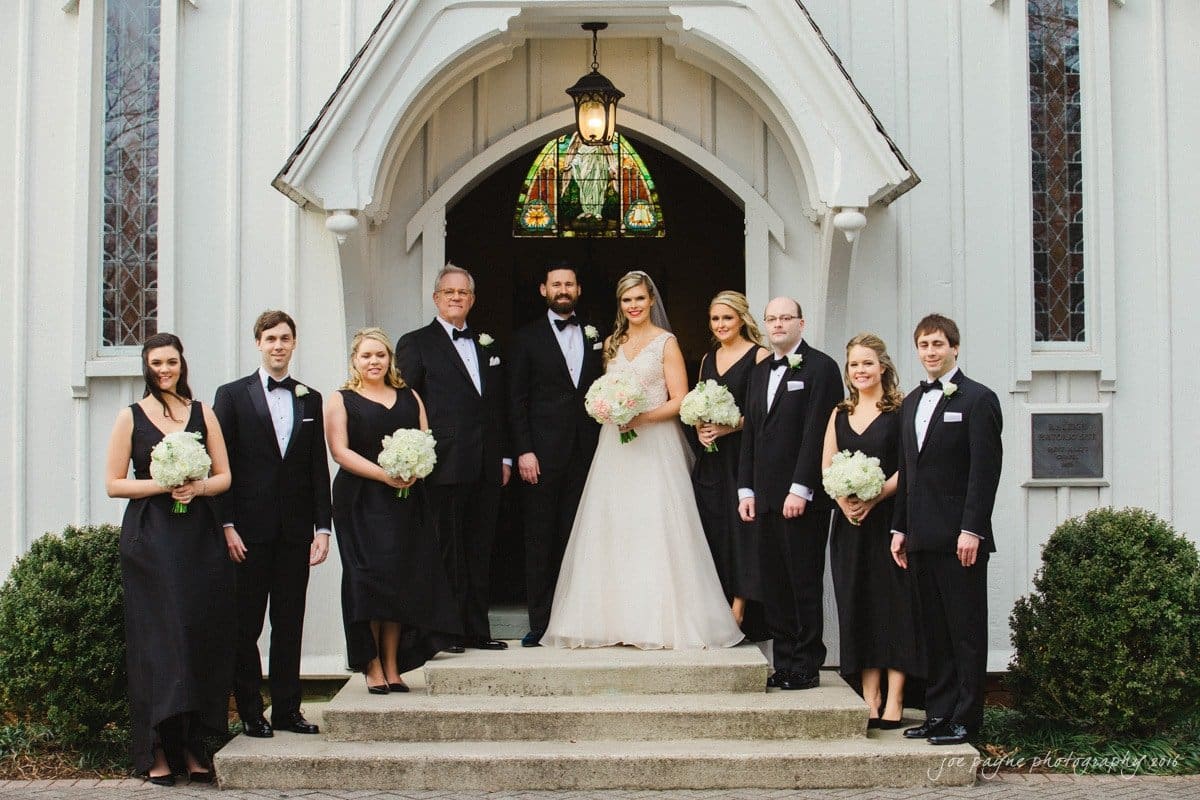st. mary's chapel raleigh wedding photography - jackie & rob A wedding party poses in front of a church entrance. The bride and groom stand in the center, surrounded by bridesmaids in black dresses and groomsmen in black suits. They are holding white bouquets and smiling. The church has a stained glass lamp above the door.