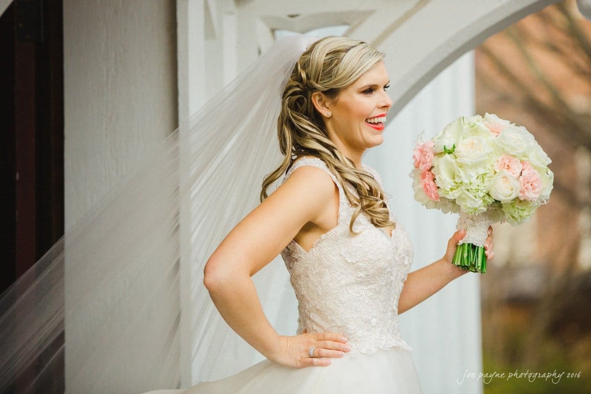 st. mary's chapel raleigh wedding photography - jackie & rob A smiling bride holding a bouquet of white and pink flowers stands under an archway. She is wearing a white lace wedding dress with a long veil, and her hair is styled in loose curls.