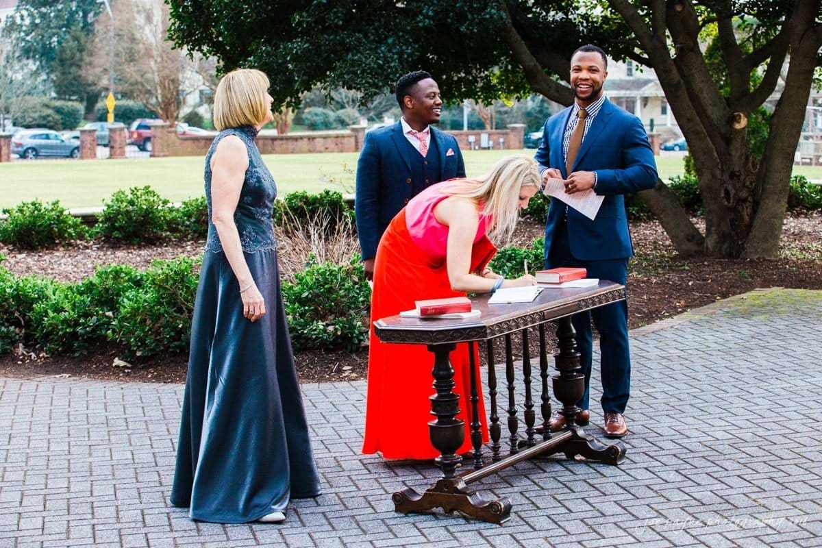 st. mary's chapel raleigh wedding photography - jackie & rob A woman in a red dress signs a book on a small table outdoors. Two men in suits and a woman in a blue dress stand nearby, smiling. They appear to be at a formal event in a garden setting.