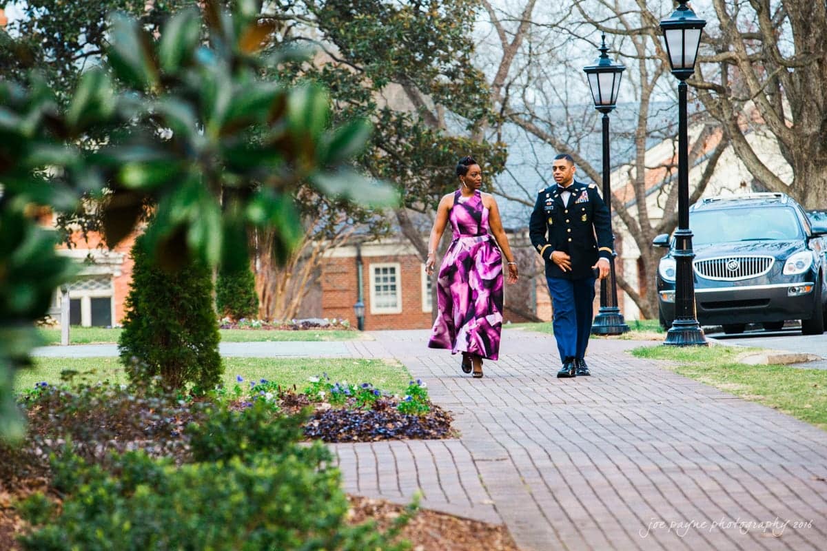 st. mary's chapel raleigh wedding photography - jackie & rob A woman in a pink and purple dress walks alongside a man in a military uniform on a brick path lined with trees and flowers. A car is parked nearby, and there are street lamps along the path.