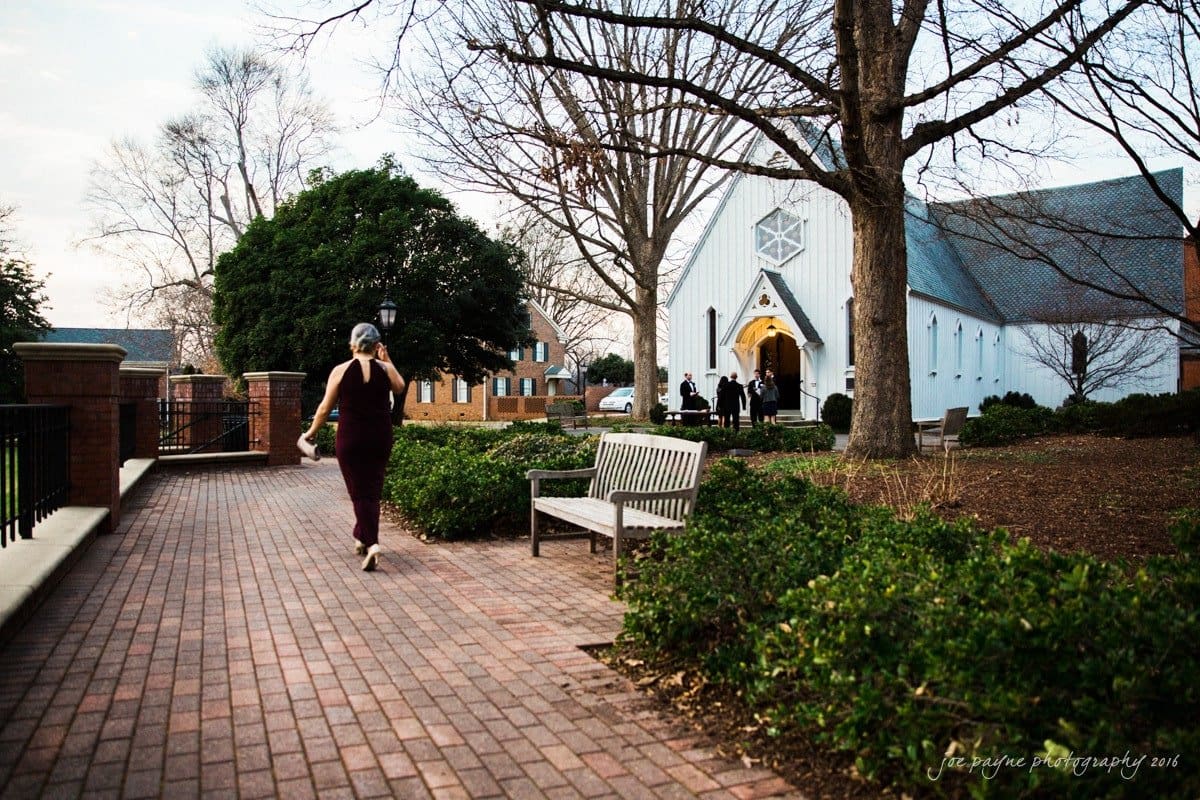 st. mary's chapel raleigh wedding photography - jackie & rob A person in a dark outfit walks on a brick path toward a white church with a lit entrance. Bare trees and a bench line the path. It is evening, and a group of people stands near the church door.