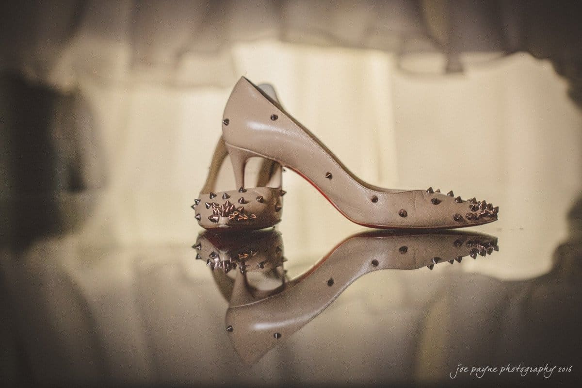 st. mary's chapel raleigh wedding photography - jackie & rob A pair of beige high-heeled shoes with spikes sit on a reflective surface, casting a mirrored image beneath them. The background is softly blurred, creating an elegant, moody atmosphere.