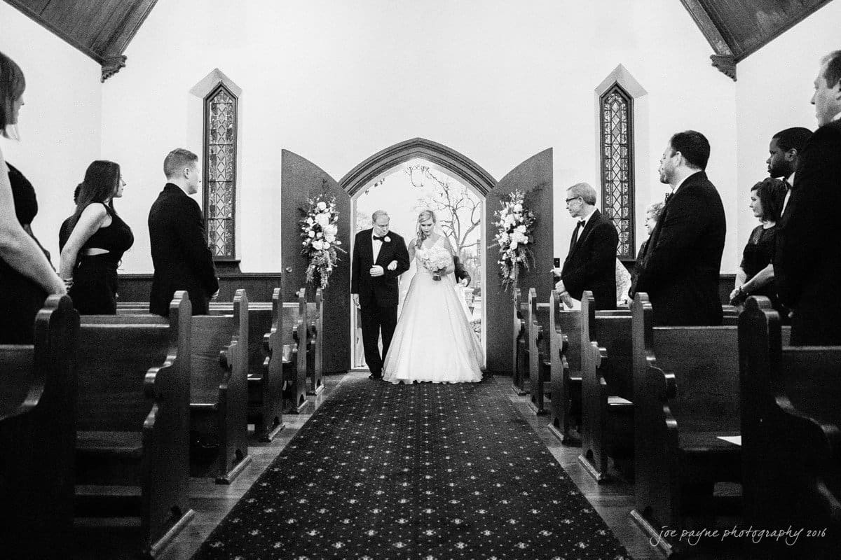 st. mary's chapel raleigh wedding photography - jackie & rob A bride walks down the aisle arm-in-arm with an older man in a church, surrounded by standing guests. The aisle is flanked by wooden pews, and two large floral arrangements stand by the open doors. The scene is captured in black and white.