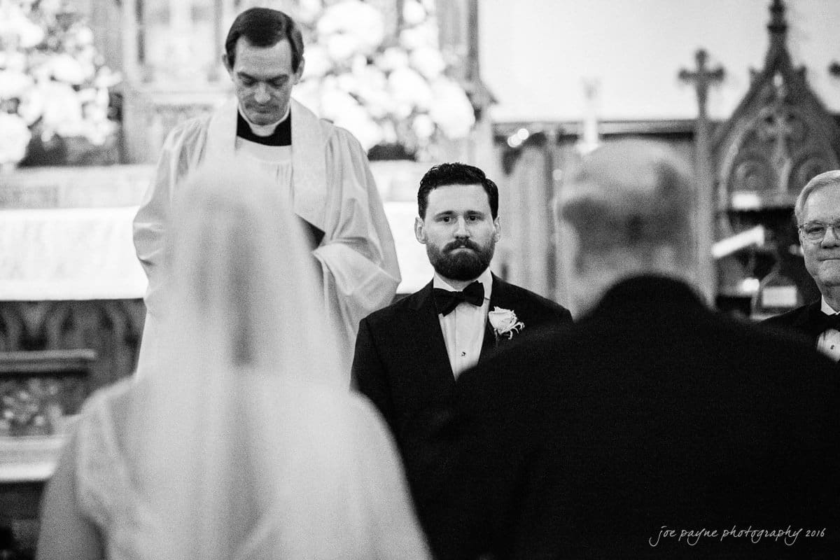 st. mary's chapel raleigh wedding photography - jackie & rob A groom stands at the altar facing the blurred figures of a bride and another person in the foreground. A clergyman stands behind the groom, with floral decorations and church elements in the background. The image is in black and white.