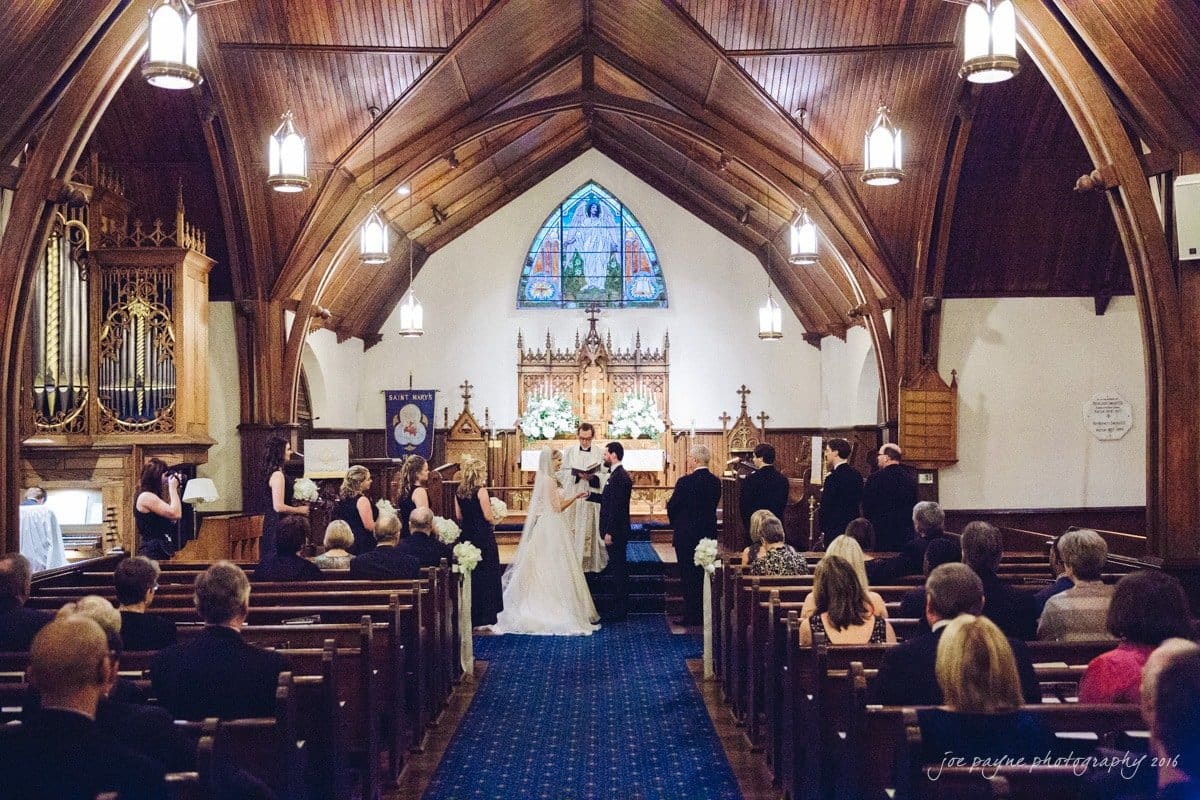 st. mary's chapel raleigh wedding photography - jackie & rob A bride and groom stand at the altar in a wooden church with a high arched ceiling. Guests are seated in pews, and the wedding party stands nearby. A stained glass window and church organ are visible in the background.