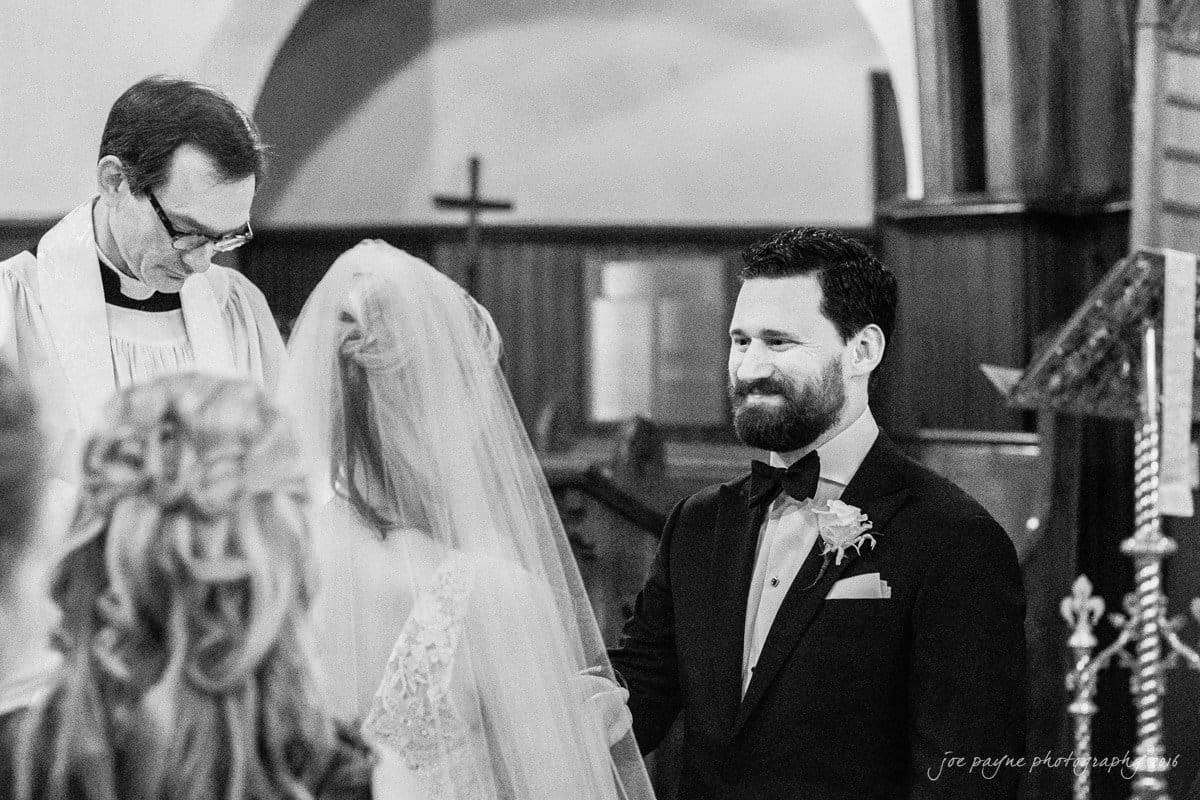 st. mary's chapel raleigh wedding photography - jackie & rob A black-and-white image of a wedding ceremony. A smiling groom in a tuxedo stands across from the bride, whose back is visible. A clergyman stands to the side. The setting is a church with a wooden interior and decorative elements.