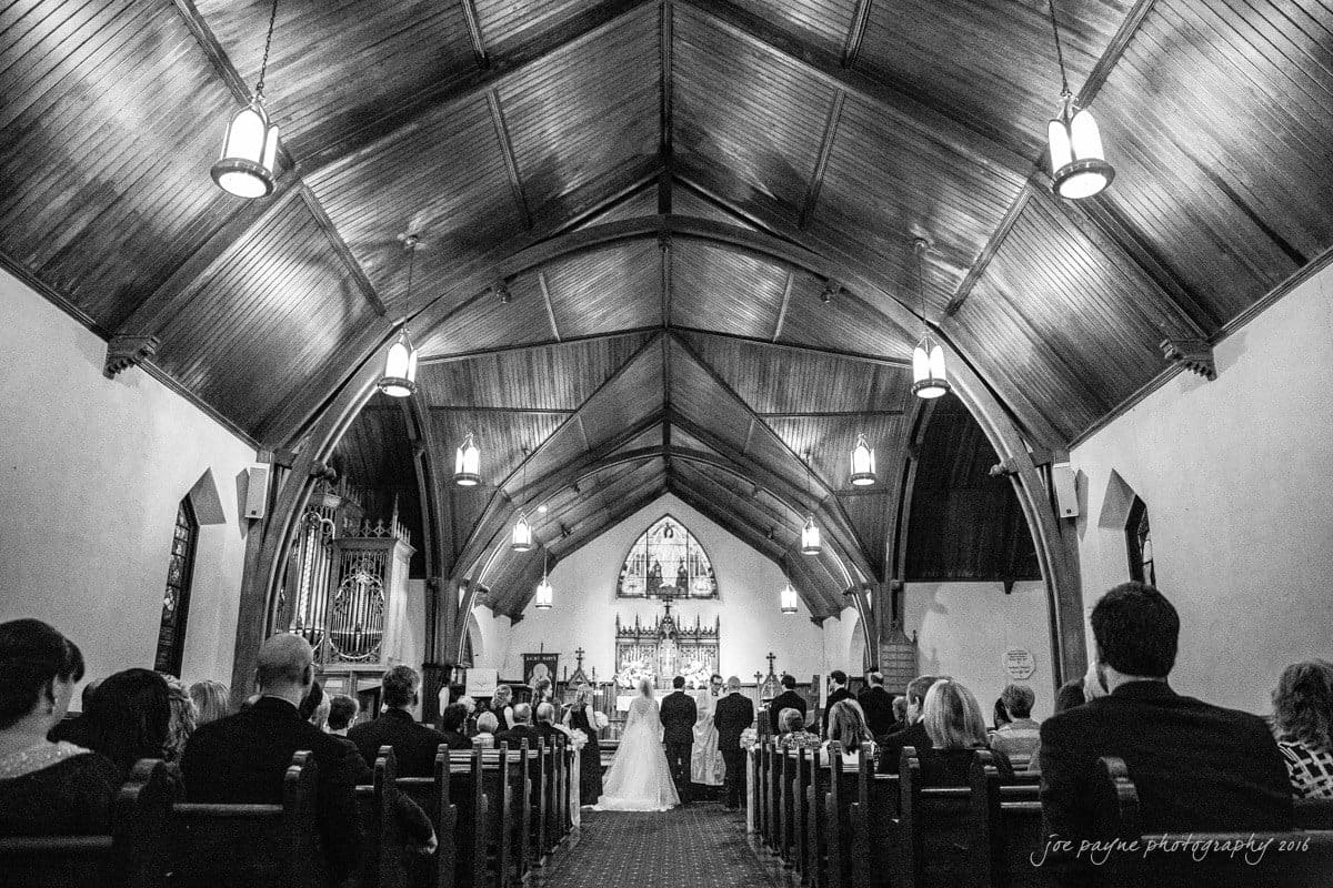 st. mary's chapel raleigh wedding photography - jackie & rob Black and white photo of a church interior during a wedding ceremony. The bride and groom stand at the altar with guests seated on either side in wooden pews, beneath a vaulted wooden ceiling with hanging lights.