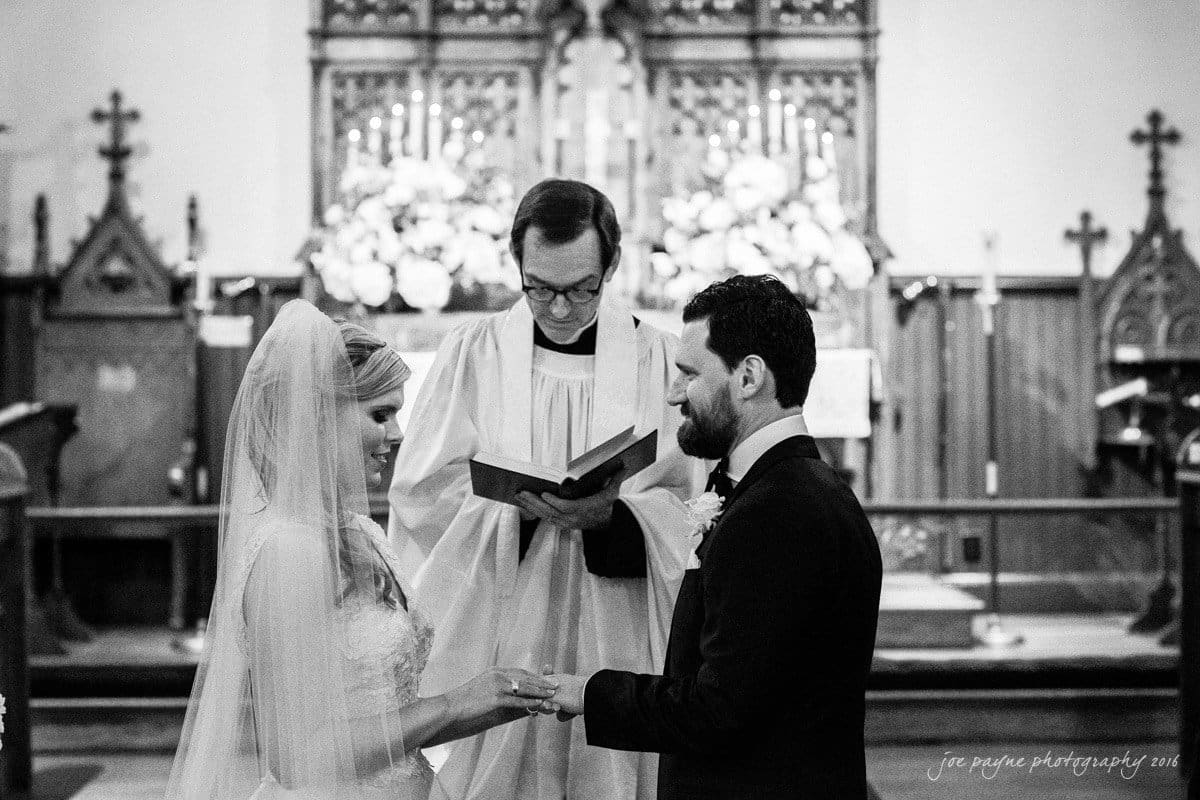 st. mary's chapel raleigh wedding photography - jackie & rob A black-and-white photo of a wedding ceremony in a church. A bride and groom stand before a minister, exchanging rings. The minister holds an open book, and the altar is adorned with flowers in the background.