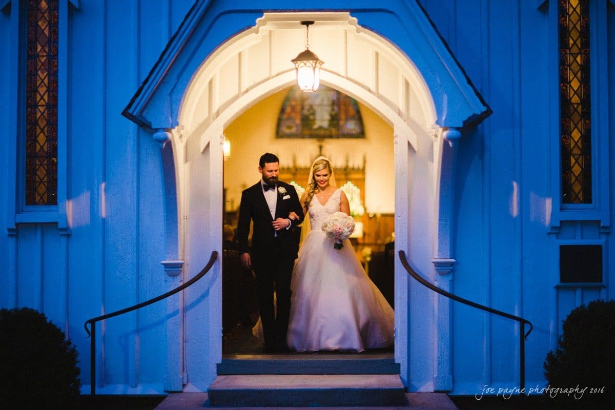 st. mary's chapel raleigh wedding photography - jackie & rob A bride in a white gown and groom in a dark tuxedo walk arm in arm under an arched doorway of a warmly lit church. The scene is illuminated by a glowing lantern above them, with the church interior softly visible.