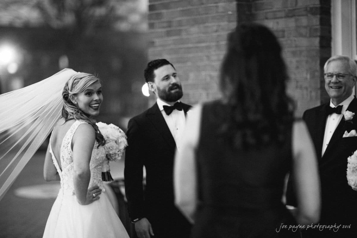 st. mary's chapel raleigh wedding photography - jackie & rob A bride in a white gown and veil smiles while holding a bouquet. A groom in a tuxedo stands beside her, looking at a woman in front of them. Another man in a tuxedo is nearby. The scene is outdoors with a brick wall in the background.