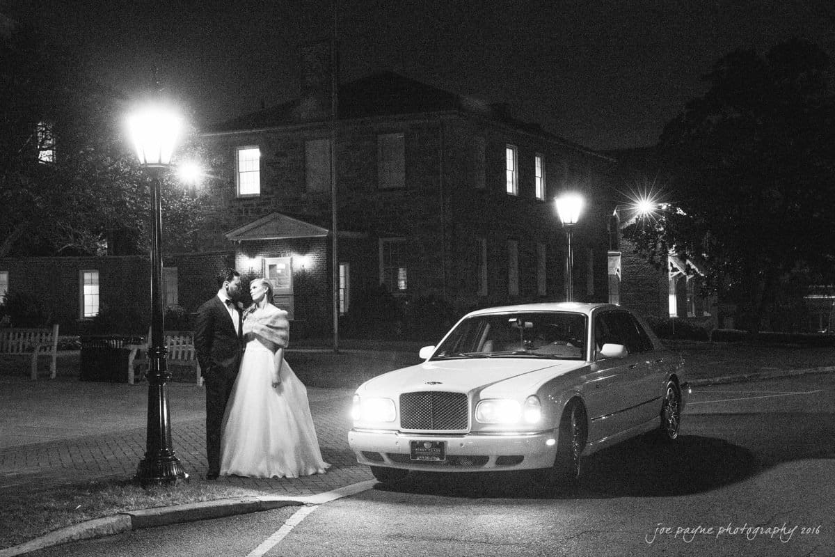 st. mary's chapel raleigh wedding photography - jackie & rob A bride and groom stand under a streetlamp at night, sharing a moment beside a vintage car. The scene is illuminated by the streetlights, with a building in the background adding to the romantic atmosphere.