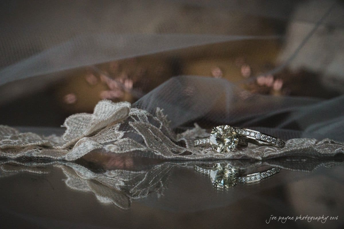 st. mary's chapel raleigh wedding photography - jackie & rob A sparkling engagement ring with a round diamond is placed on delicate lace fabric. The scene is softly lit, casting a reflection of the ring on a glass surface underneath.