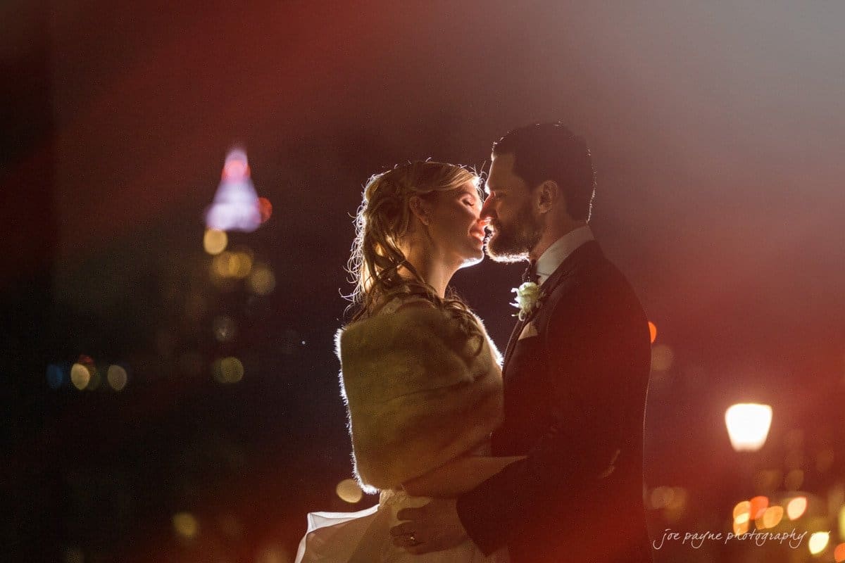st. mary's chapel raleigh wedding photography - jackie & rob A couple in formal attire shares an intimate moment outdoors at night. The background is softly lit with city lights, including a recognizable illuminated building. The scene is warm and romantic, with a dreamy, soft-focus effect.