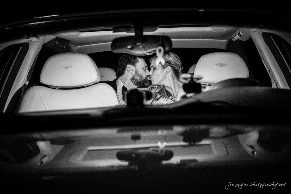 st. mary's chapel raleigh wedding photography - jackie & rob A couple sits in the front seats of a car, leaning in for a kiss. The photo is in black and white, taken from the outside through the windshield. The car interior is visible, and they appear to be dressed for a formal event.
