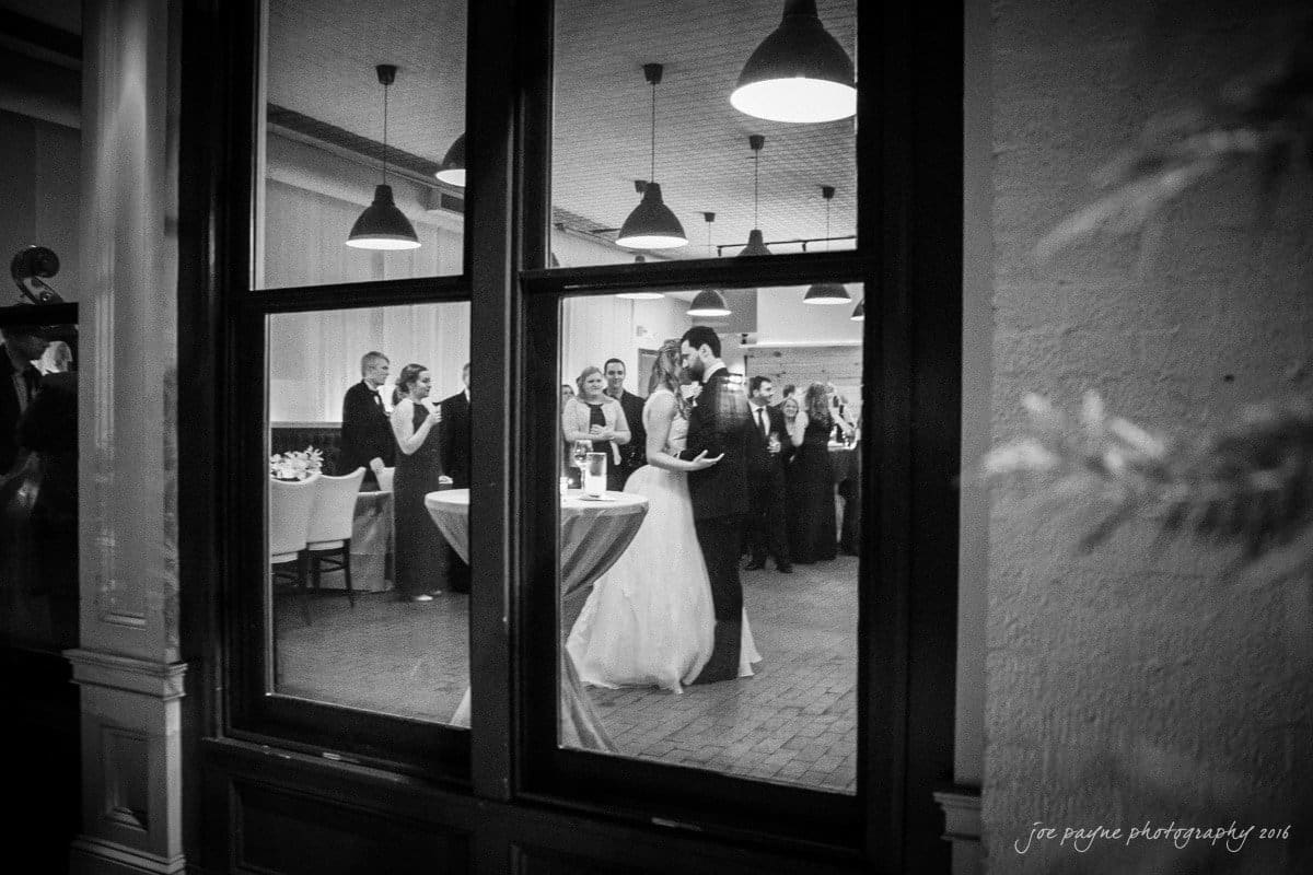 st. mary's chapel raleigh wedding photography - jackie & rob A black-and-white photo showing a couple dancing inside a venue, viewed through a window. Guests are standing around elegant tables, some holding drinks. Overhead lights illuminate the scene, creating a warm, festive atmosphere.
