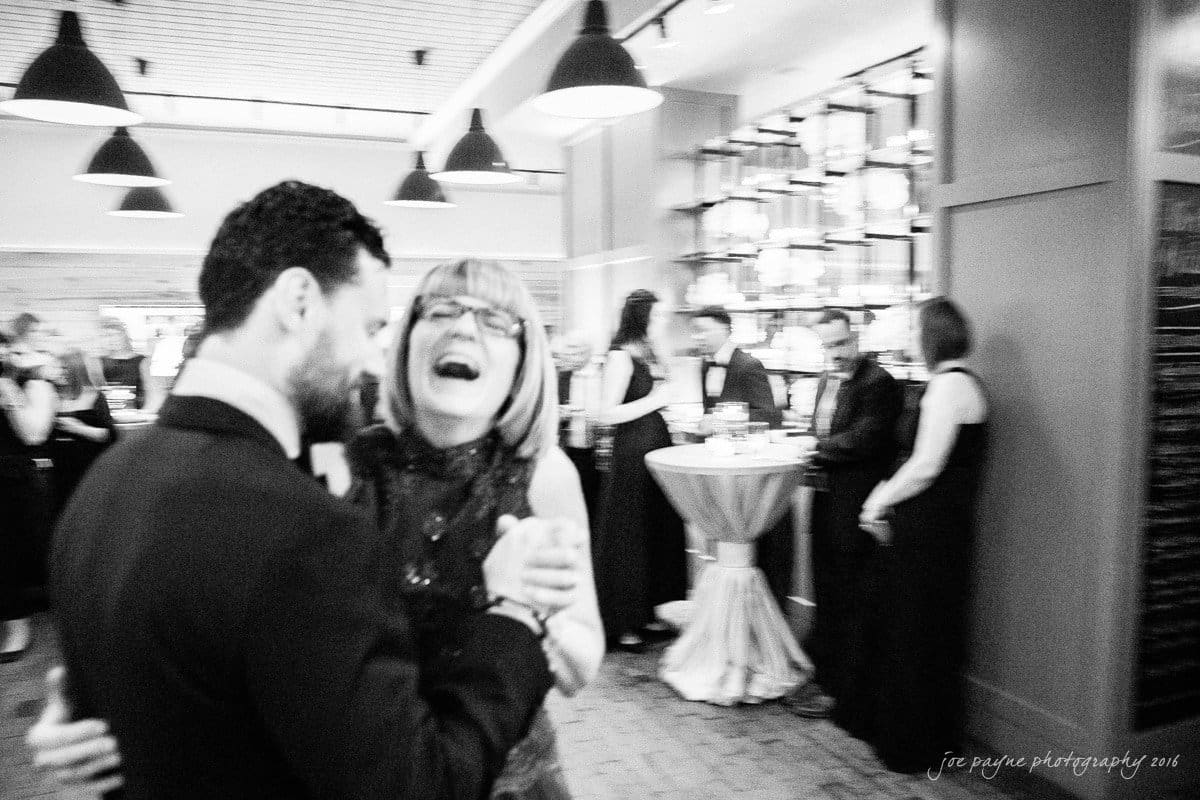 st. mary's chapel raleigh wedding photography - jackie & rob A black and white photo of a couple laughing and dancing in a lively setting. Other elegantly dressed people are mingling and socializing around a bar area in the background.