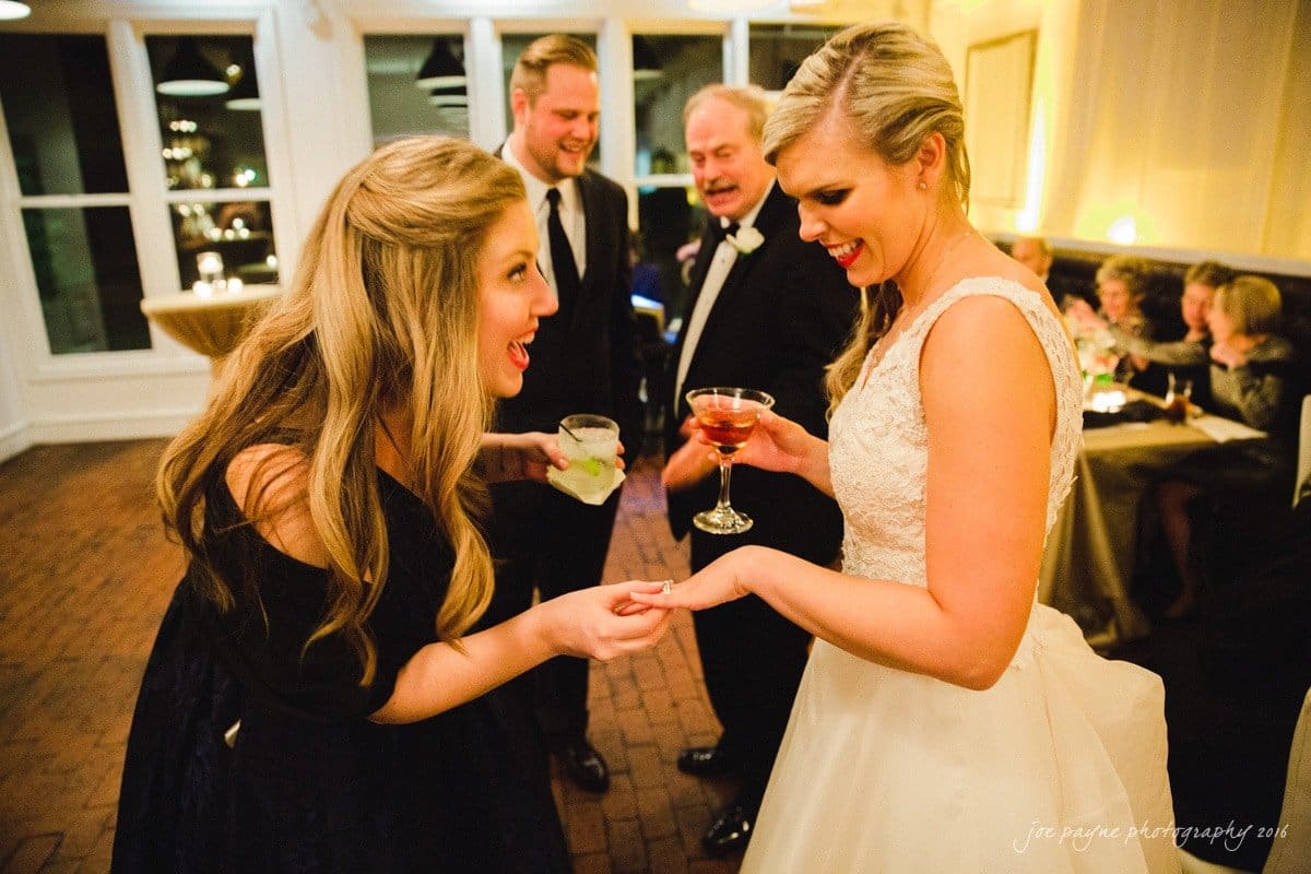 st. mary's chapel raleigh wedding photography - jackie & rob A bride in a white dress shows her ring to a smiling woman holding a drink. Two men, one in a suit and another in a tuxedo, stand nearby in a warmly lit room with other guests in the background.