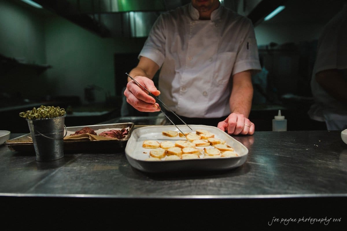 st. mary's chapel raleigh wedding photography - jackie & rob A chef in a white uniform carefully arranges food on a large rectangular plate in a dimly lit kitchen. The counter is set with various ingredients and tools.