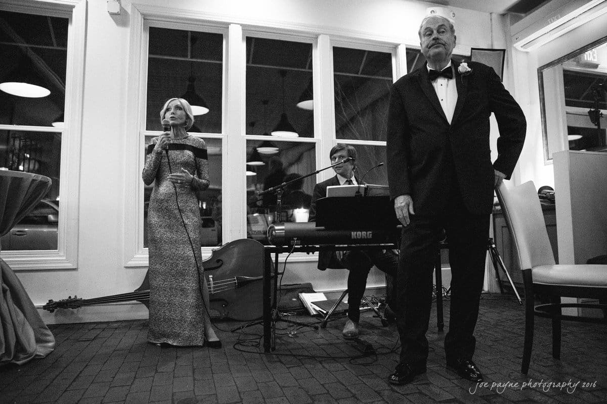 st. mary's chapel raleigh wedding photography - jackie & rob A black and white photo of a formal event with a woman singing into a microphone, a man playing the keyboard, and another man in a tuxedo standing nearby. A double bass leans against the wall in the background.