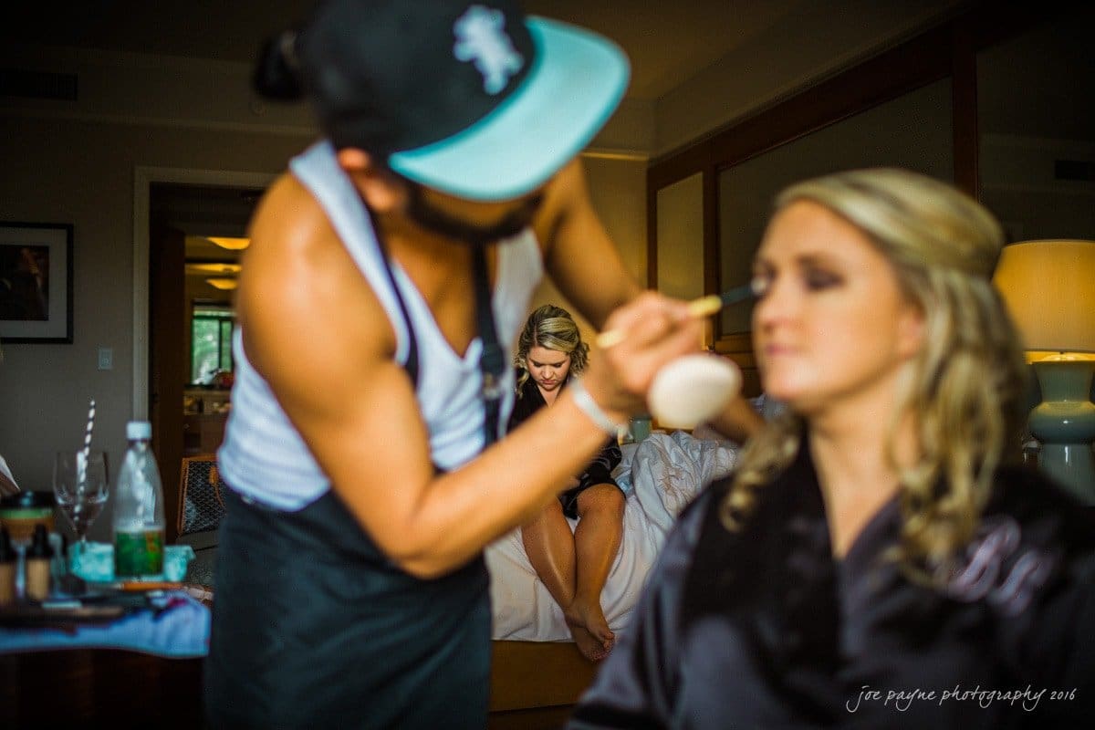 st. mary's chapel raleigh wedding photography - jackie & rob A makeup artist applies foundation to a woman seated in a room, while another woman sits on a bed in the background. The room is softly lit, with makeup supplies visible on a nearby surface.