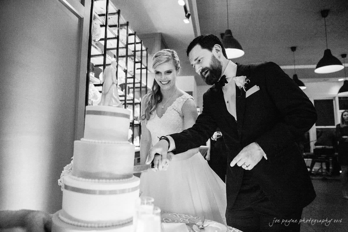 st. mary's chapel raleigh wedding photography - jackie & rob A bride and groom smile as they cut a tiered wedding cake together in a festive indoor setting. The bride holds the knife while the groom assists. The room is decorated with lights and guests are visible in the background.