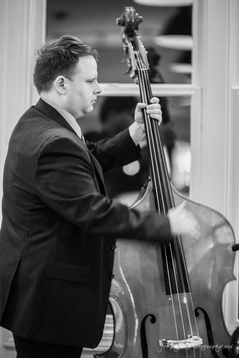 st. mary's chapel raleigh wedding photography - jackie & rob A man in a dark suit plays an upright bass indoors. The image is in black and white, capturing his focused expression as he stands near a window.