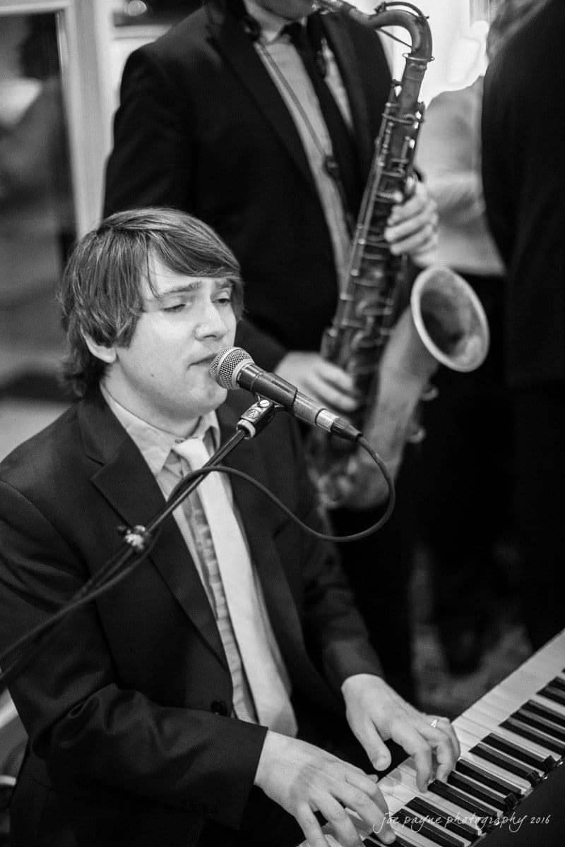st. mary's chapel raleigh wedding photography - jackie & rob A black and white photo of a musician playing piano and singing into a microphone. In the background, another musician plays a saxophone. Both are dressed in suits, and the setting appears to be a live performance.