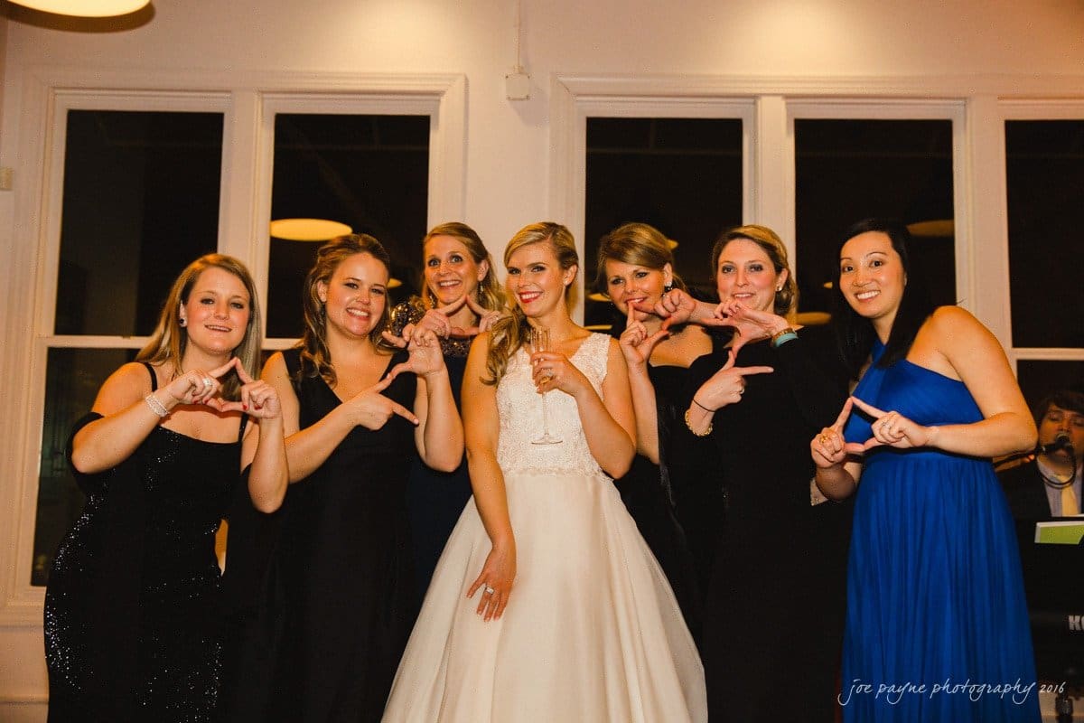 st. mary's chapel raleigh wedding photography - jackie & rob A bride in a white gown stands with six women in formal dresses at an indoor event. Some of them are making hand signs. There are large windows in the background.