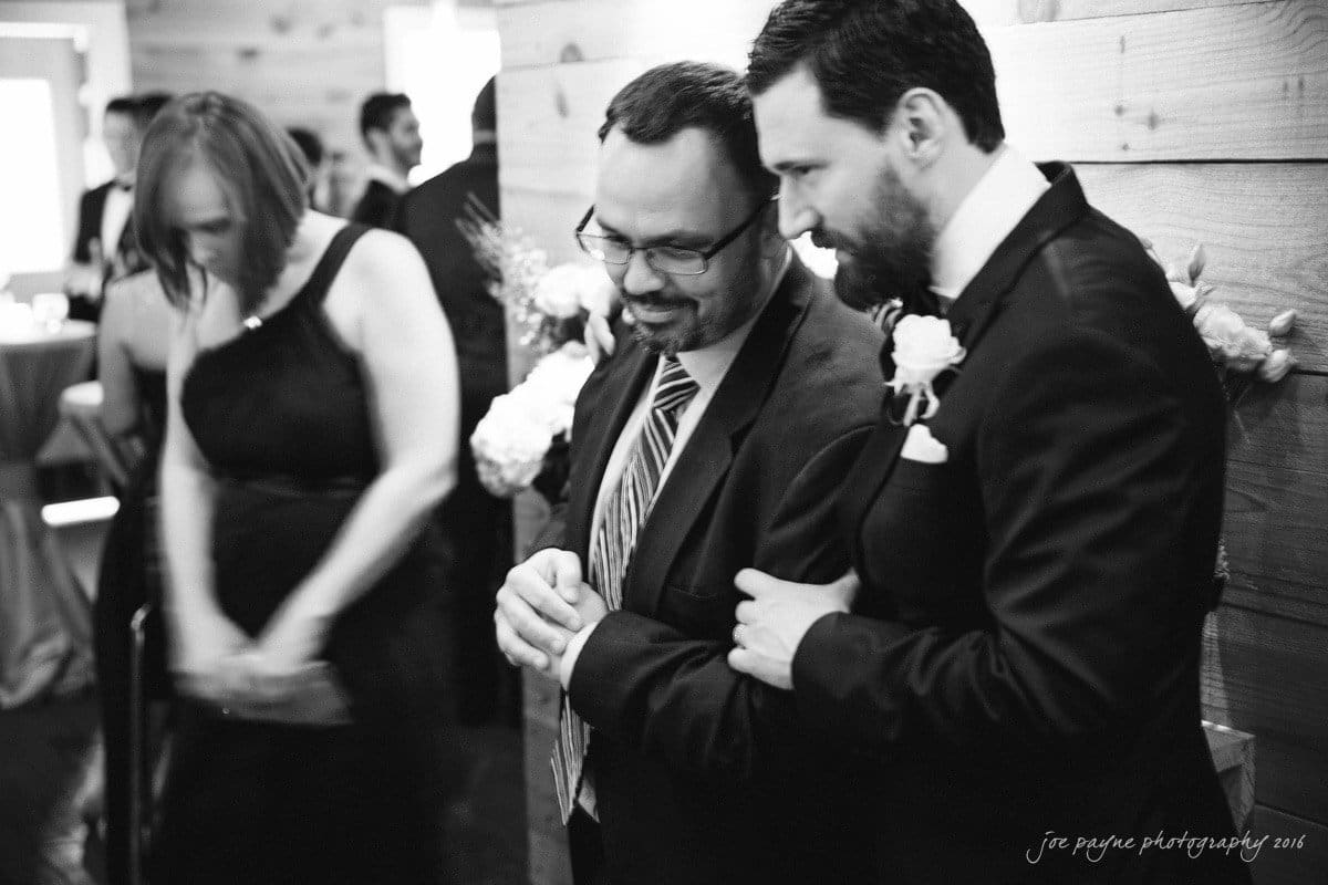 st. mary's chapel raleigh wedding photography - jackie & rob A black and white photo shows two men in suits, one with a boutonniere, standing and smiling. A woman in a dress is blurred in motion on the left. The setting appears to be a formal indoor event, with wooden walls and floral decorations.