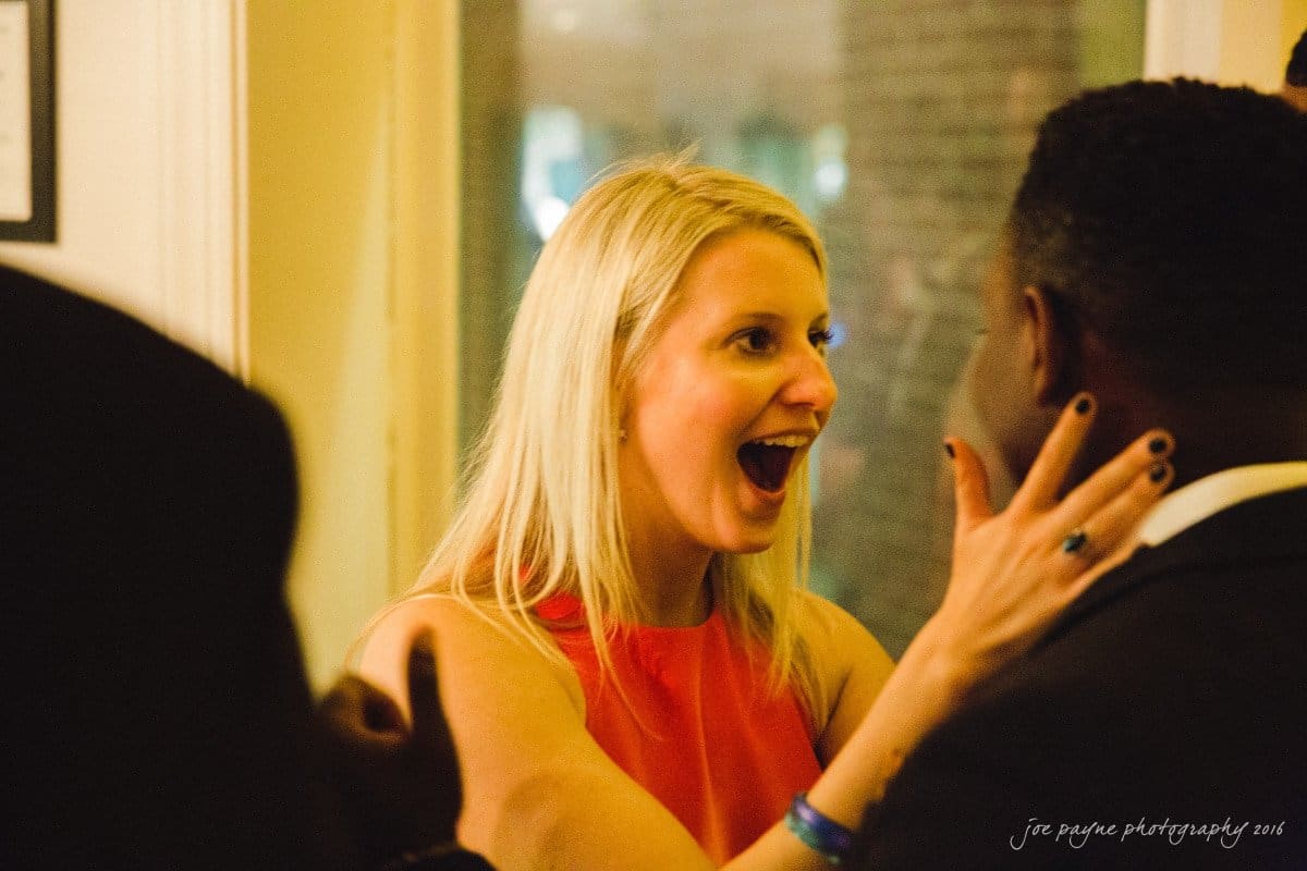 st. mary's chapel raleigh wedding photography - jackie & rob A woman in a red dress smiles widely with open arms facing a man in a dark suit. The setting appears to be indoors, with warm lighting.