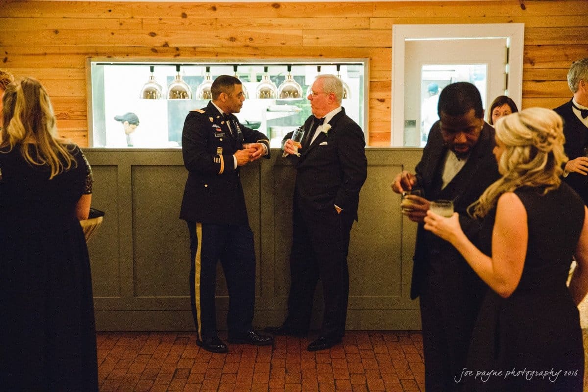st. mary's chapel raleigh wedding photography - jackie & rob People are gathered in an elegantly decorated room. Two men, one in a military uniform and the other in a suit, converse by a counter. Others chat and hold drinks. The setting exudes a formal and relaxed atmosphere.