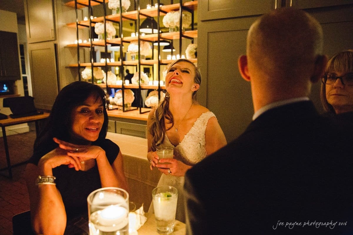 st. mary's chapel raleigh wedding photography - jackie & rob A bride in a white dress is joyfully smiling and tilting her head back at a dinner table. Three other people are seated nearby, engaged in conversation. The setting is warmly lit with candles and decor in the background.