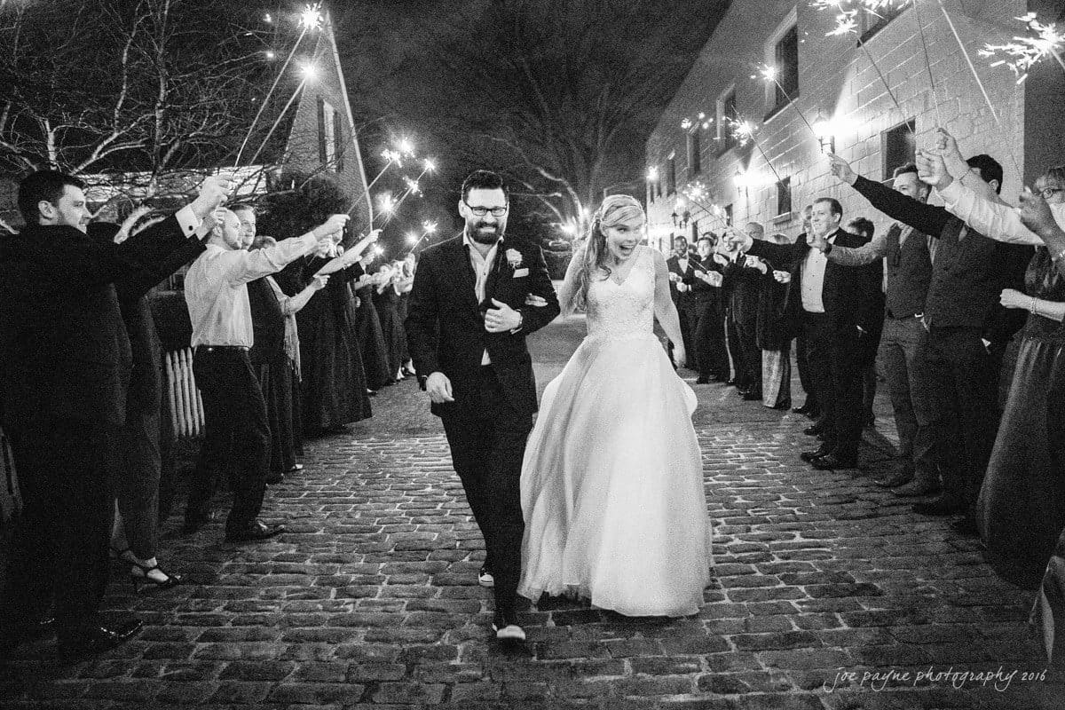 st. mary's chapel raleigh wedding photography - jackie & rob A newlywed couple walks down a cobblestone path at night, smiling, while guests hold sparklers aloft on either side, creating a festive atmosphere. The bride wears a long white gown and the groom is in a suit.