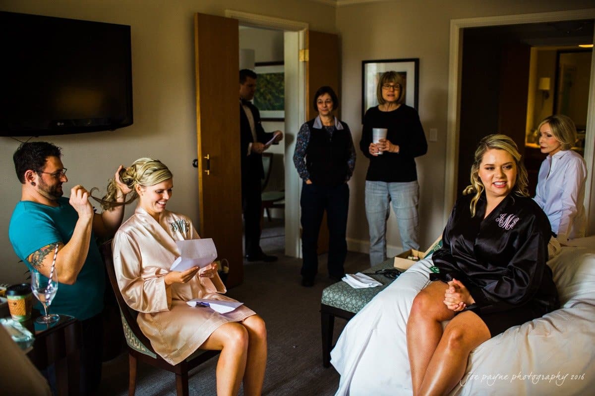 st. mary's chapel raleigh wedding photography - jackie & rob A group of people in a room, with two women seated and others standing. The seated women are wearing robes and one is having her hair styled. Some are smiling and holding drinks, suggesting a festive or preparatory gathering, possibly before an event.