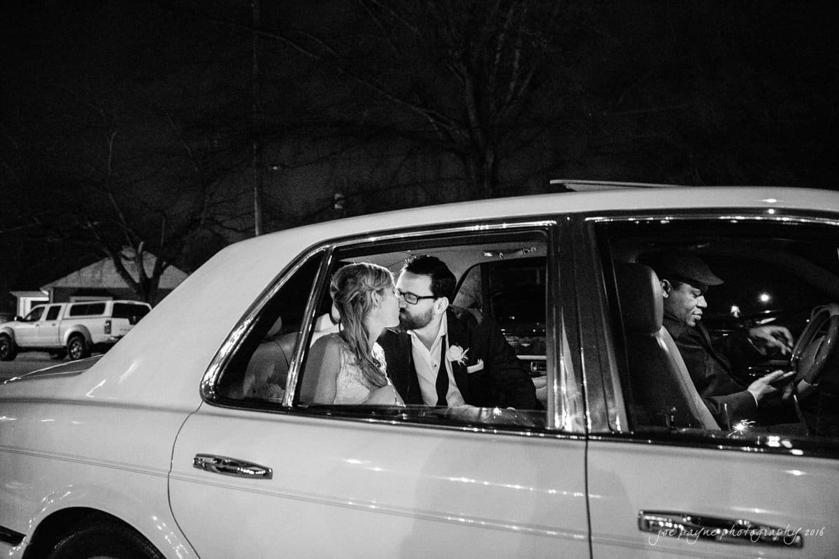 st. mary's chapel raleigh wedding photography - jackie & rob A black and white photo of a couple sitting in the backseat of a vintage car, kissing, while the driver looks forward. They are surrounded by parked cars and trees at night, creating a romantic atmosphere.