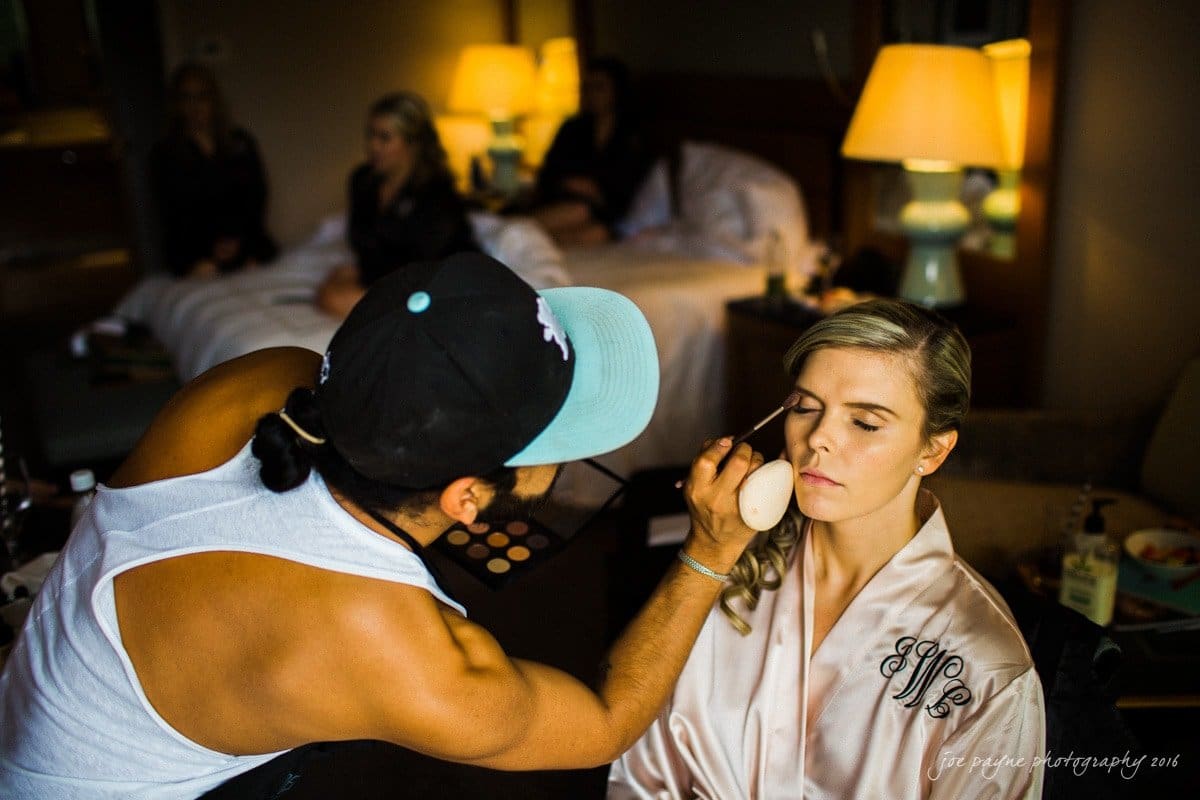st. mary's chapel raleigh wedding photography - jackie & rob A makeup artist applies cosmetics to a seated woman wearing a silk robe in a softly lit room. Two other women are blurred in the background on a bed. The atmosphere is calm and focused.