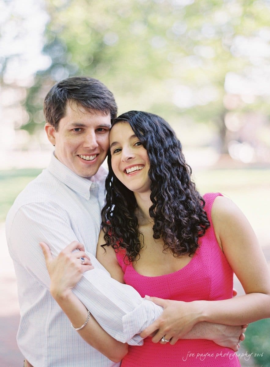 A couple smiling and embracing each other in a sunlit outdoor setting. The man is wearing a white shirt, and the woman is wearing a pink dress. Blurred trees and greenery are in the background.