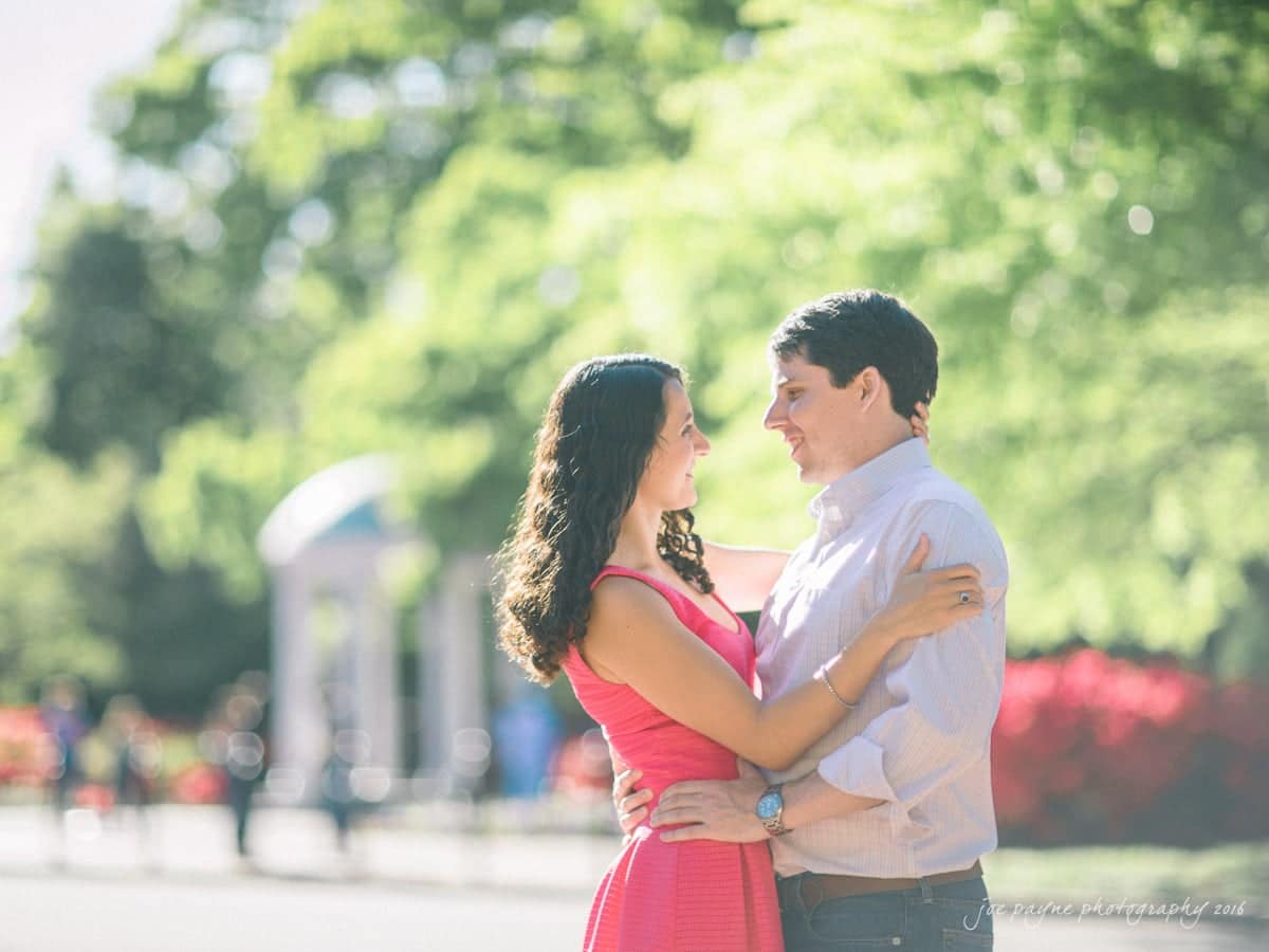 A couple stands embraced in a sunlit park, gazing into each others eyes. The woman wears a pink dress, and the man wears a light shirt. Lush greenery and a domed structure provide a scenic backdrop.