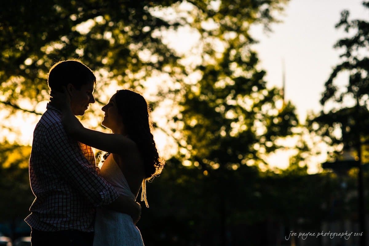 Silhouetted couple embracing in a park during sunset, with trees and soft light in the background.